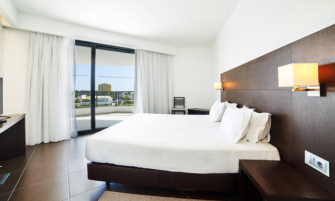 Modern resort bedroom with queen bed, brown headwall, and sliding glass doors opening to a balcony overlooking the grounds at Alvor Baía Resort Hotel.