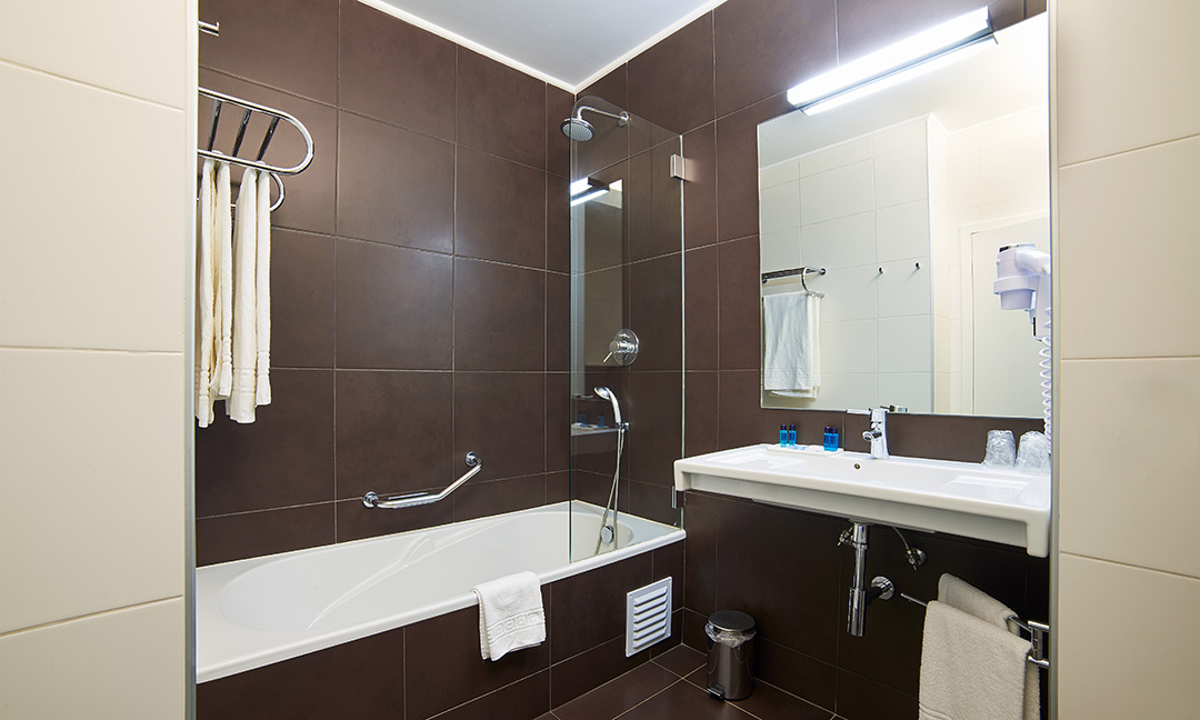 Modern bathroom with dark brown and cream tiles, soaking tub with glass shower enclosure, and vanity with large mirror at Alvor Baía Resort Hotel.