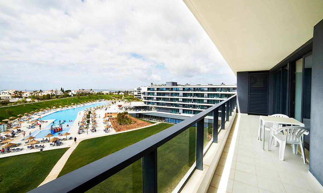 View from a balcony at Alvor Baía Resort Hotel overlooking a large swimming pool, manicured lawns, and multi-story resort buildings with a coastal town in the distance.