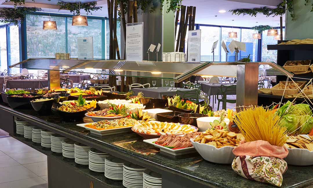 Buffet spread at Alvor Baía Resort Hotel with salads, pasta, meats, and bread displayed under heat lamps along a dark counter.