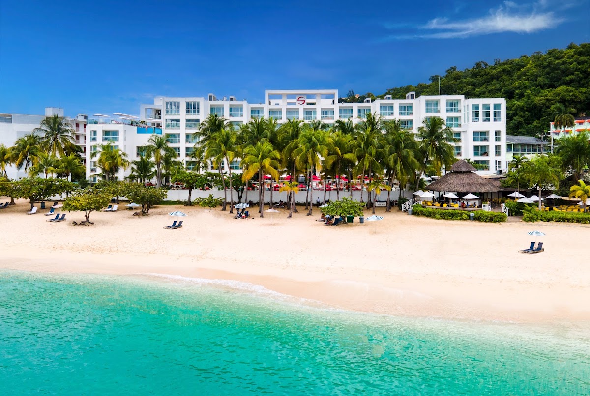 White multi-story resort building with turquoise shutters sits behind a sandy beach lined with palm trees, lounge chairs, and thatched umbrellas facing clear turquoise water.