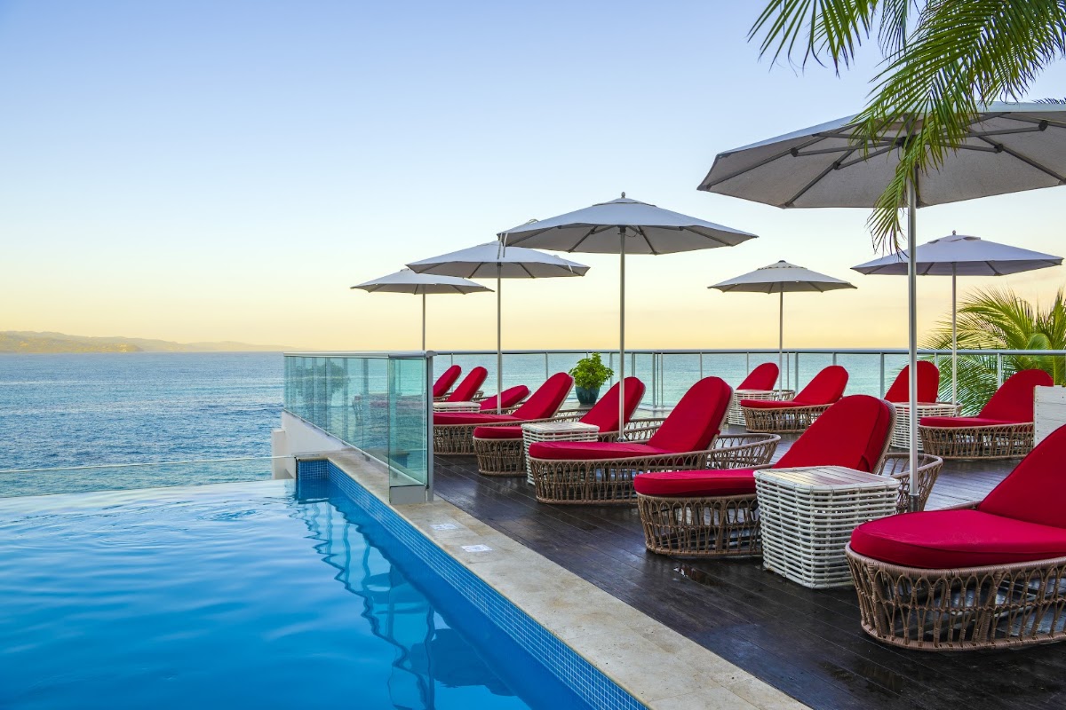 Infinity pool and lounge deck at S Hotel Montego Bay with red cushioned chairs, umbrellas, and ocean view at sunset.