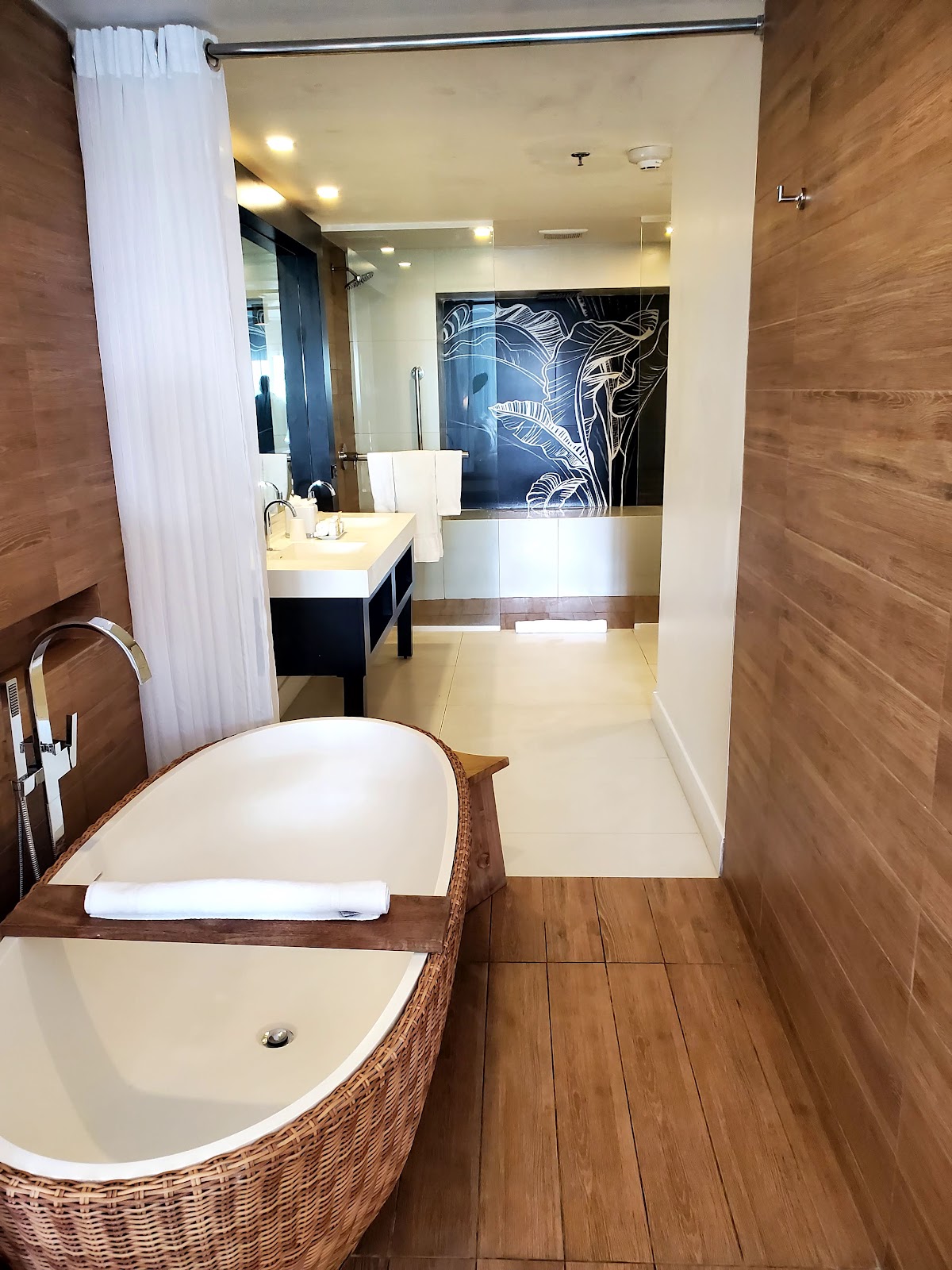 Resort bathroom with woven soaking tub in foreground, wooden flooring, and glass shower with decorative tile work beyond.
