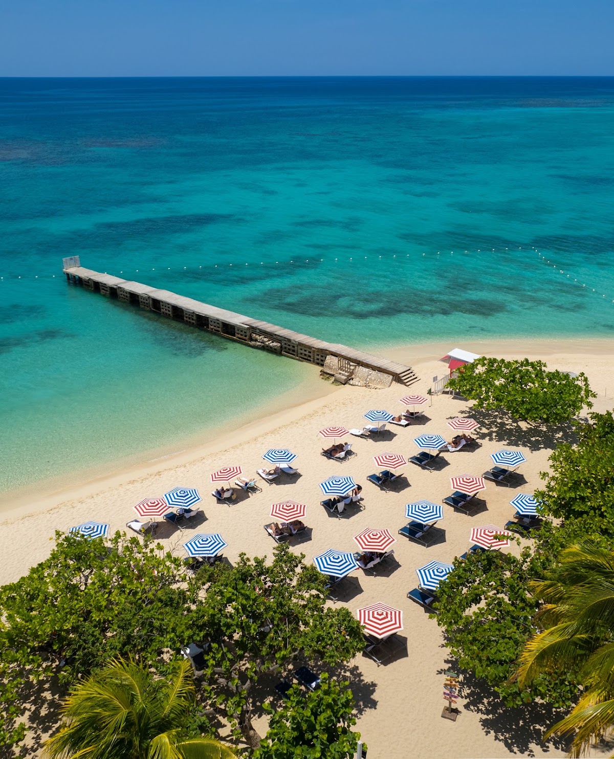 Sandy beach lined with striped umbrellas and lounge chairs, with a long pier extending into turquoise water at S Hotel Montego Bay.