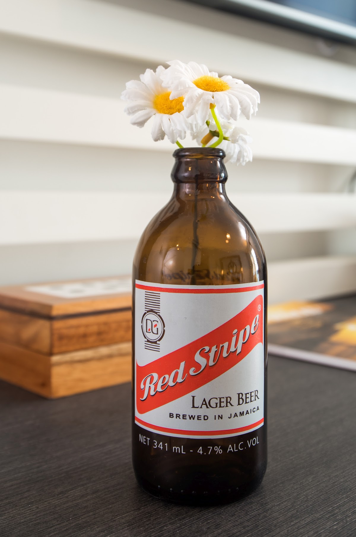 Red Stripe beer bottle with white daisies in the neck, sitting on a dark table with a wooden box and white louvered background.