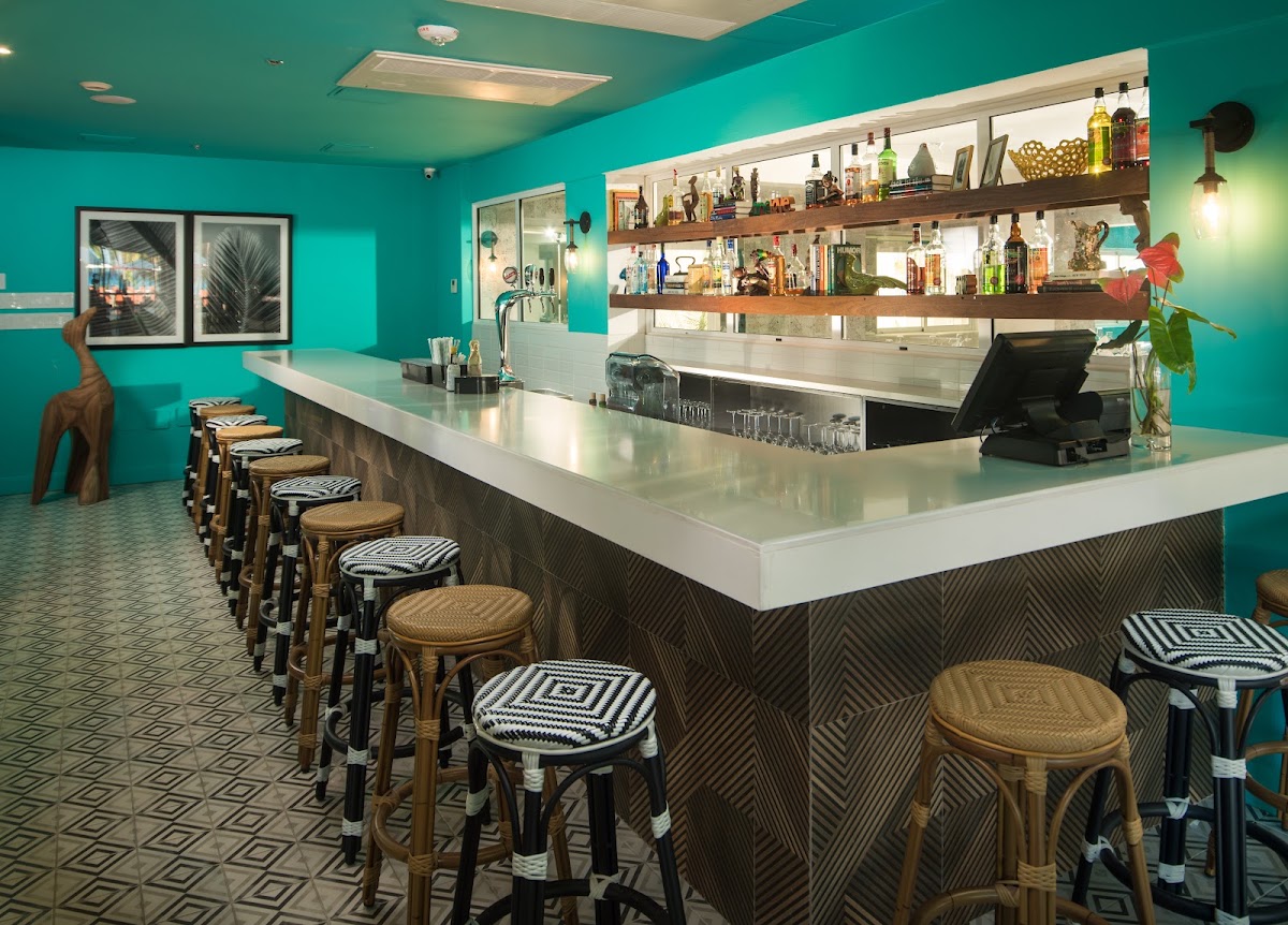 Turquoise bar with white counter, wooden stools with patterned cushions, and shelves of liquor bottles behind the counter.