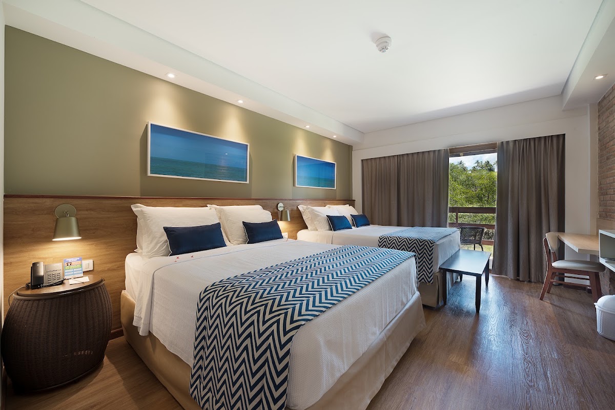 Modern resort room with two queen beds, blue and white geometric bedding, ocean-view artwork, and a balcony overlooking tropical greenery.