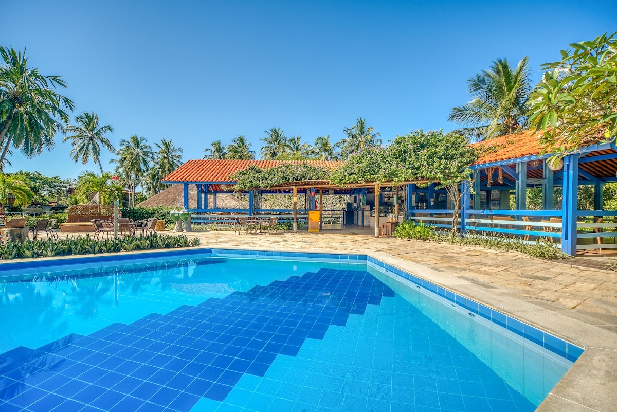 Blue swimming pool with tiled deck, palm trees, and blue-and-orange buildings with thatched roofs at Salinas Maragogi.