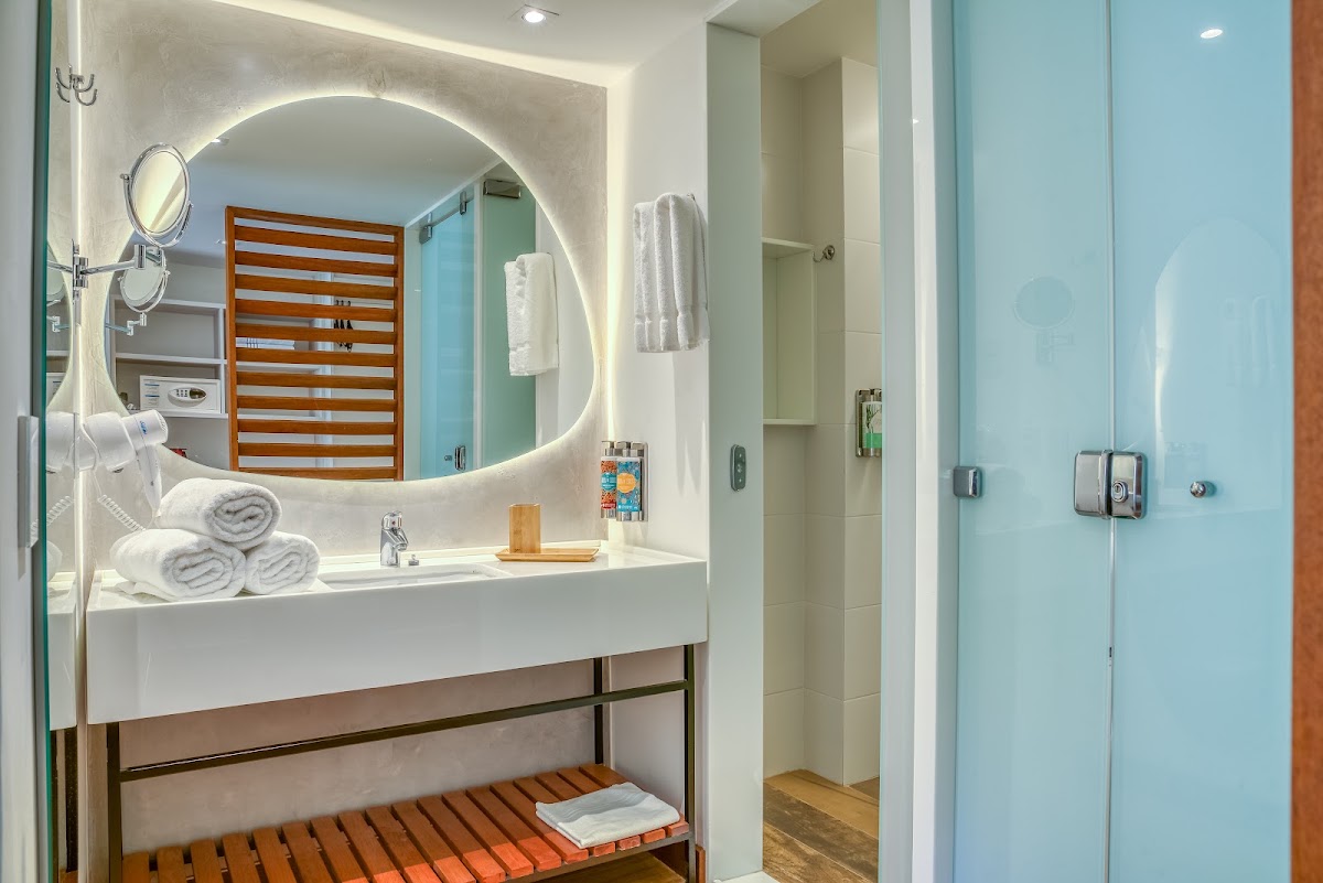 Bathroom at Salinas Maragogi with round illuminated mirror, white sink, rolled towels, wooden bench, and glass shower enclosure with light blue tile.