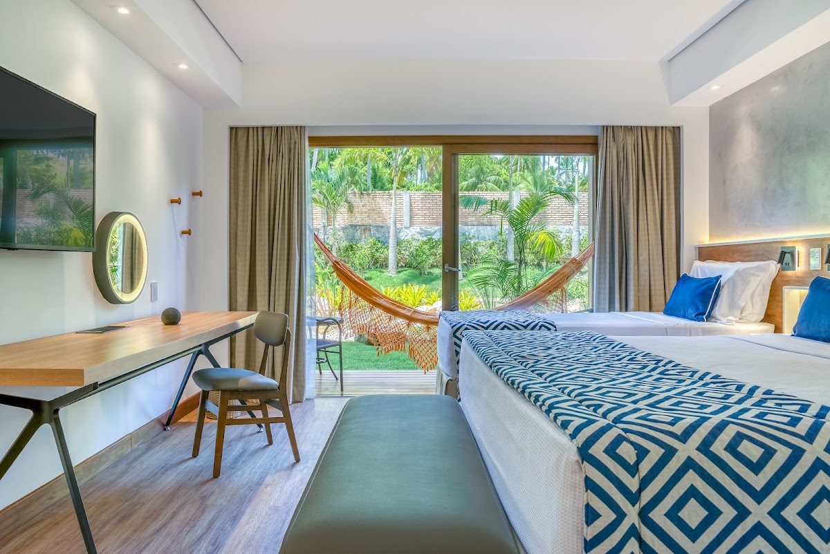 Resort bedroom with queen bed, desk, and glass doors opening to private patio with hammock and tropical garden at Salinas Maragogi.
