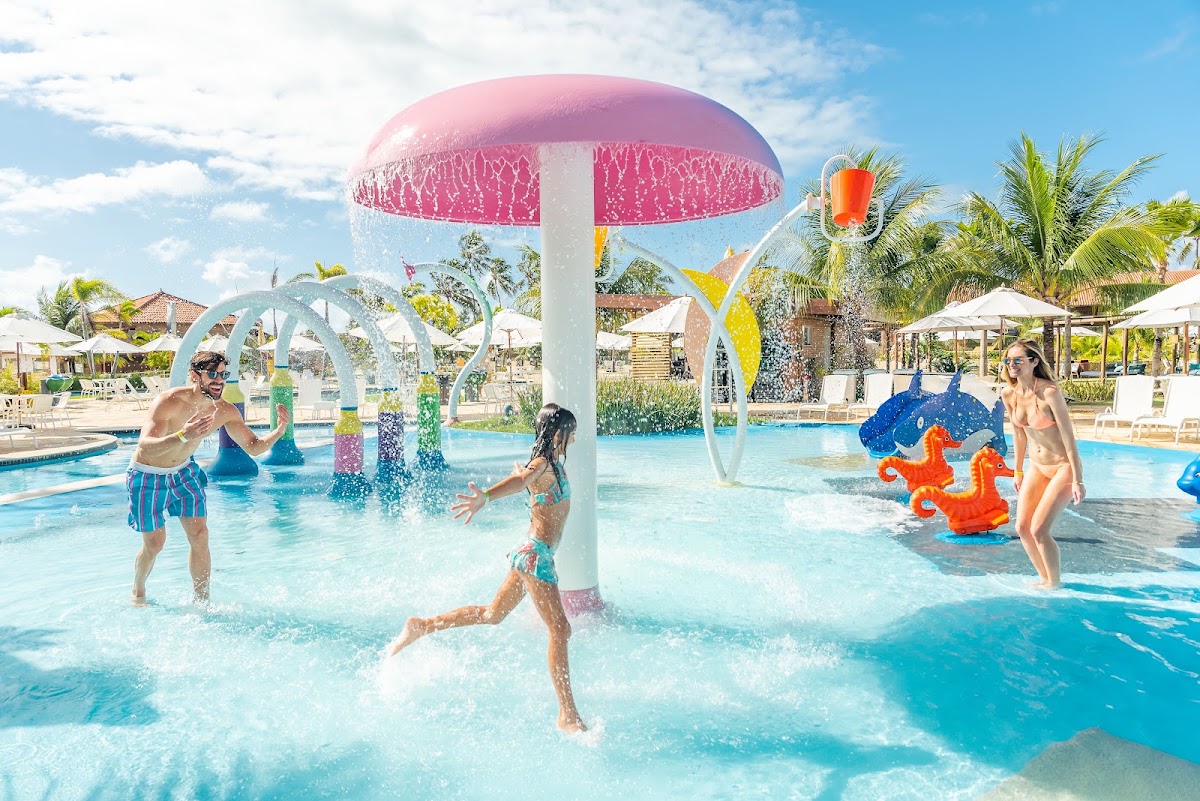 Families and children play in a shallow pool with colorful water features and a pink umbrella spray structure at Salinas Maragogi All Inclusive Resort.