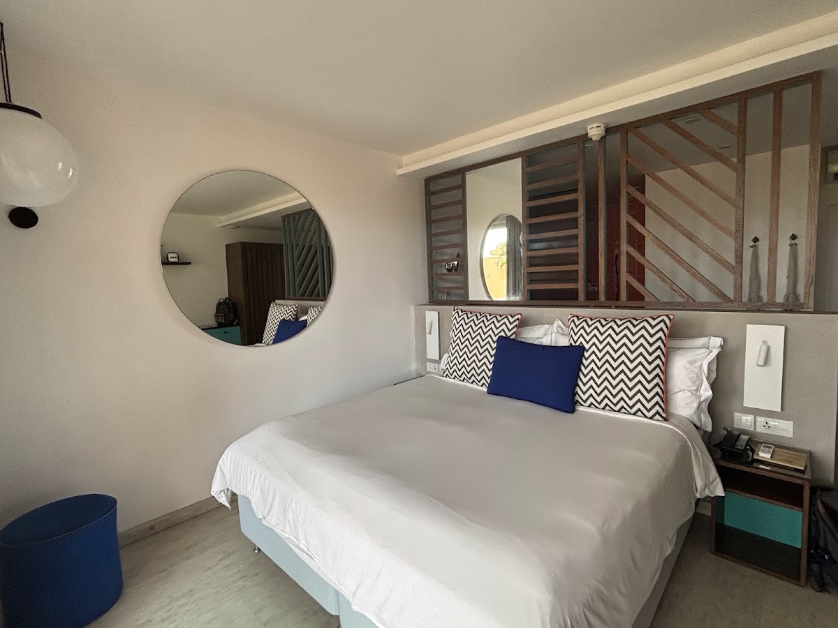 Modern resort bedroom with queen bed, geometric patterned pillows, round mirror, and wooden shuttered windows overlooking the ocean.