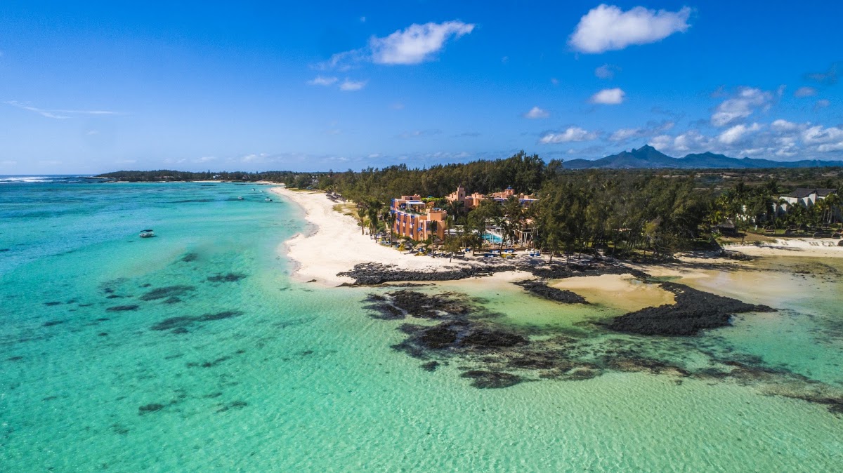 Aerial view of SALT of Palmar resort on a turquoise lagoon with white sandy beach, palm trees, and volcanic mountains in the distance.