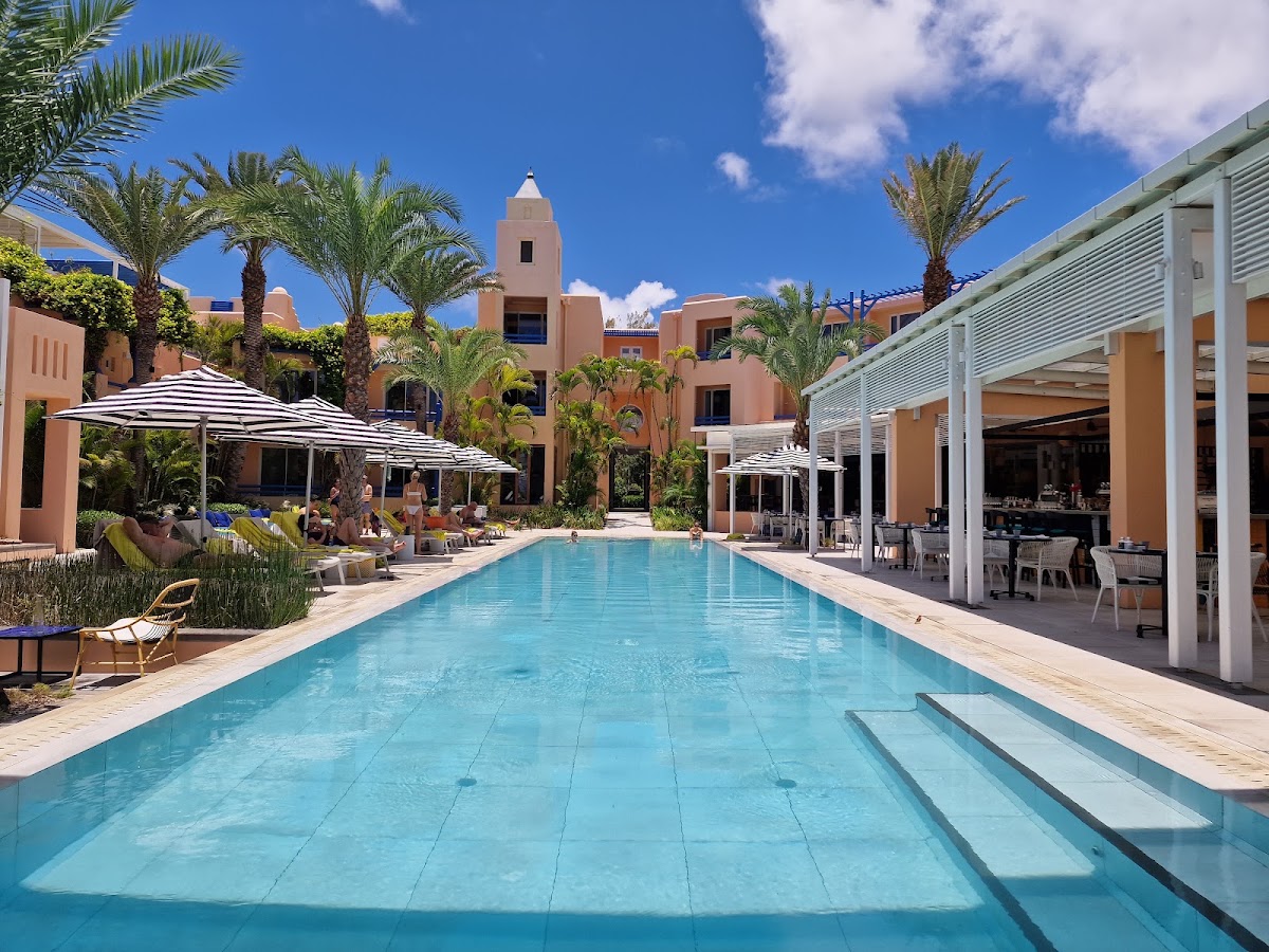 Long rectangular pool with turquoise water flanked by terracotta buildings, palm trees, striped umbrellas, and a white pergola bar area under clear blue sky.