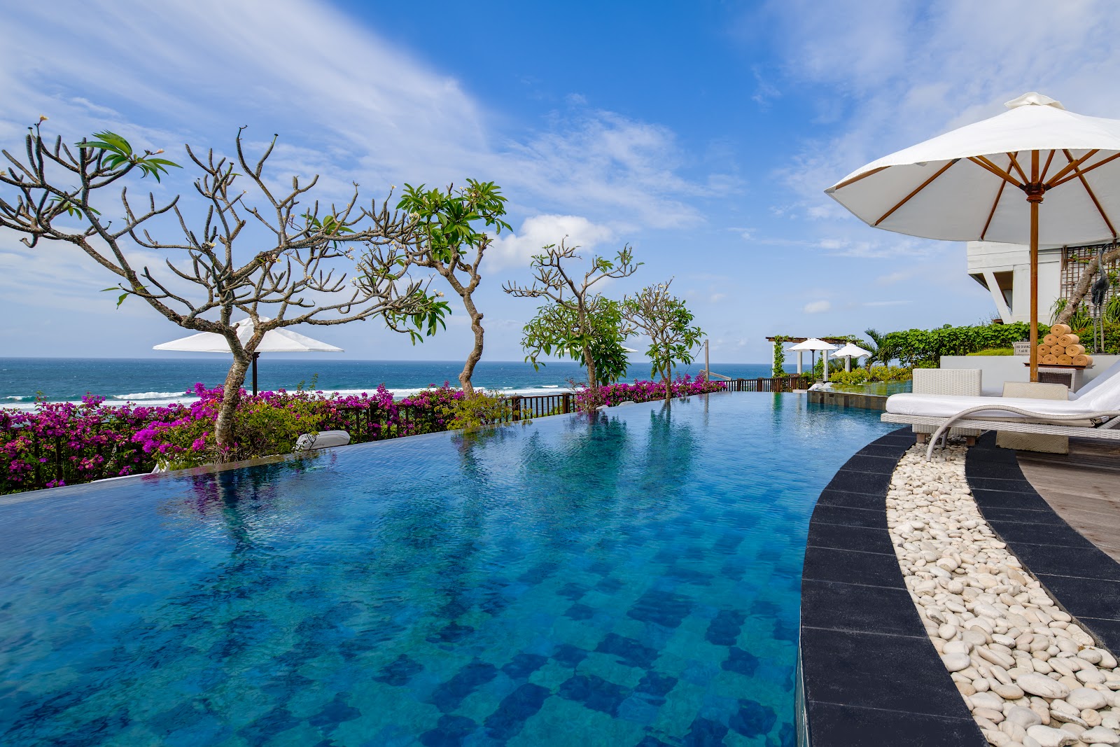 Infinity pool at Samabe Bali Suites & Villas overlooking the ocean, with magenta bougainvillea, plumeria trees, loungers, and white umbrellas.
