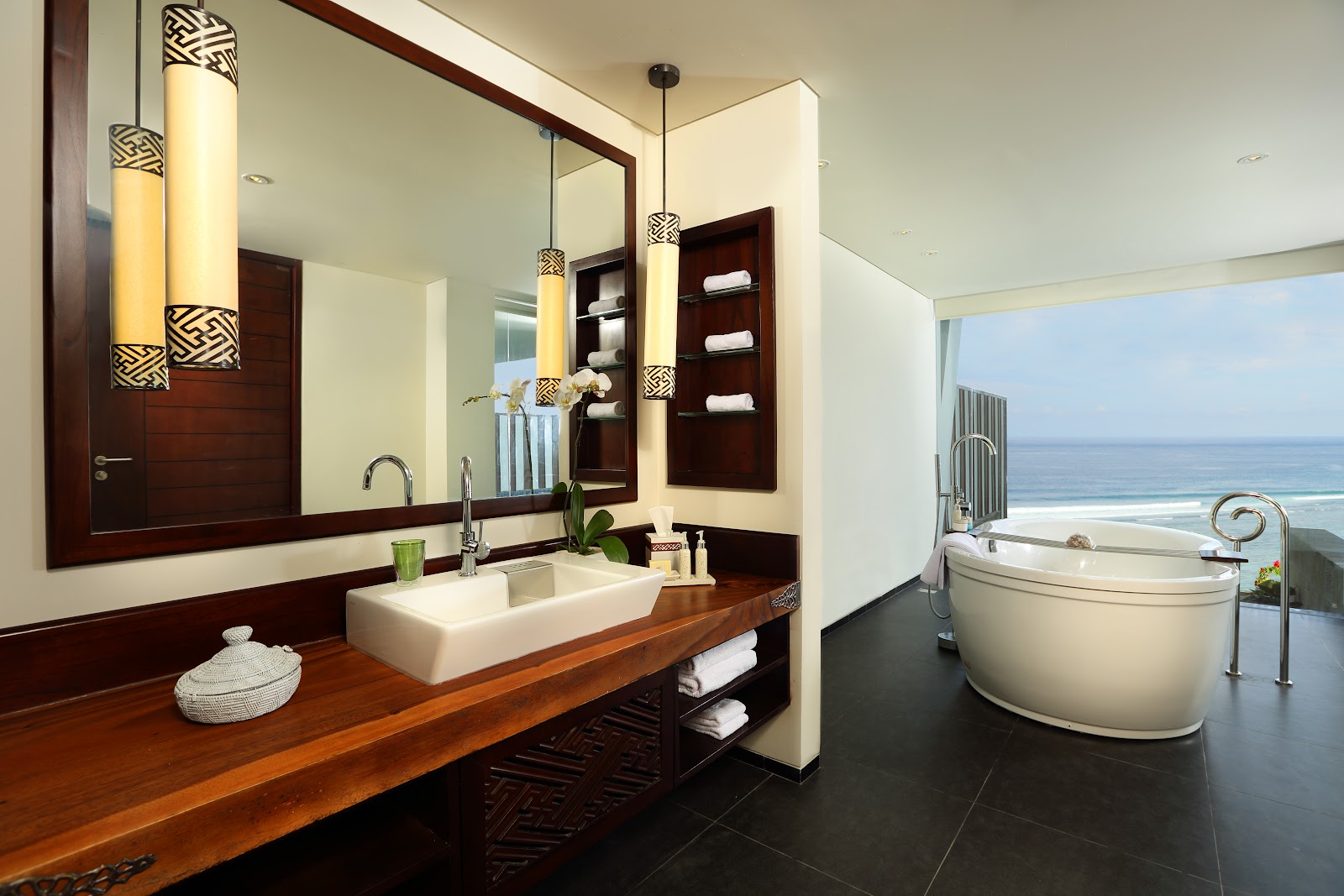 Samabe Bali Suites & Villas bathroom with dark wood vanity, white vessel sink, large mirror, pendant lights, and curved soaking tub overlooking the ocean.
