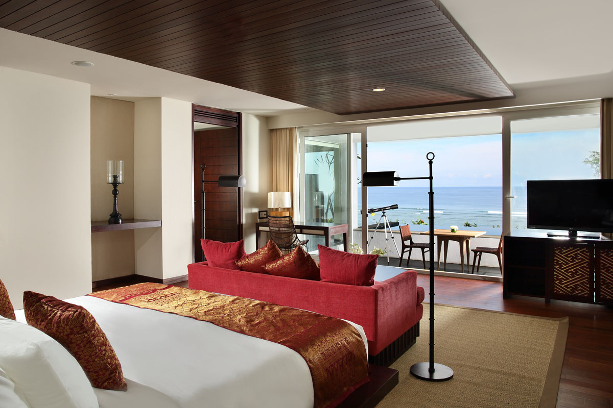 Upscale resort suite with red sofa, white bed, dark wood ceiling, and ocean views through glass doors opening to a balcony.
