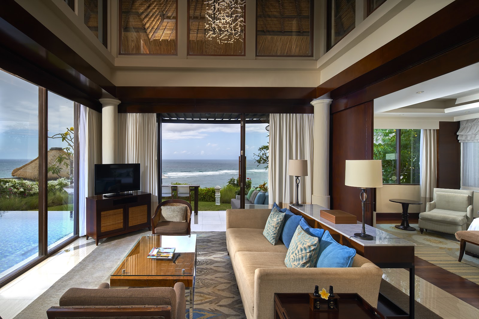 Samabe Bali Suites & Villas suite with ocean views through floor-to-ceiling windows, beige sofa, dark wood furnishings, and infinity pool visible on the left.