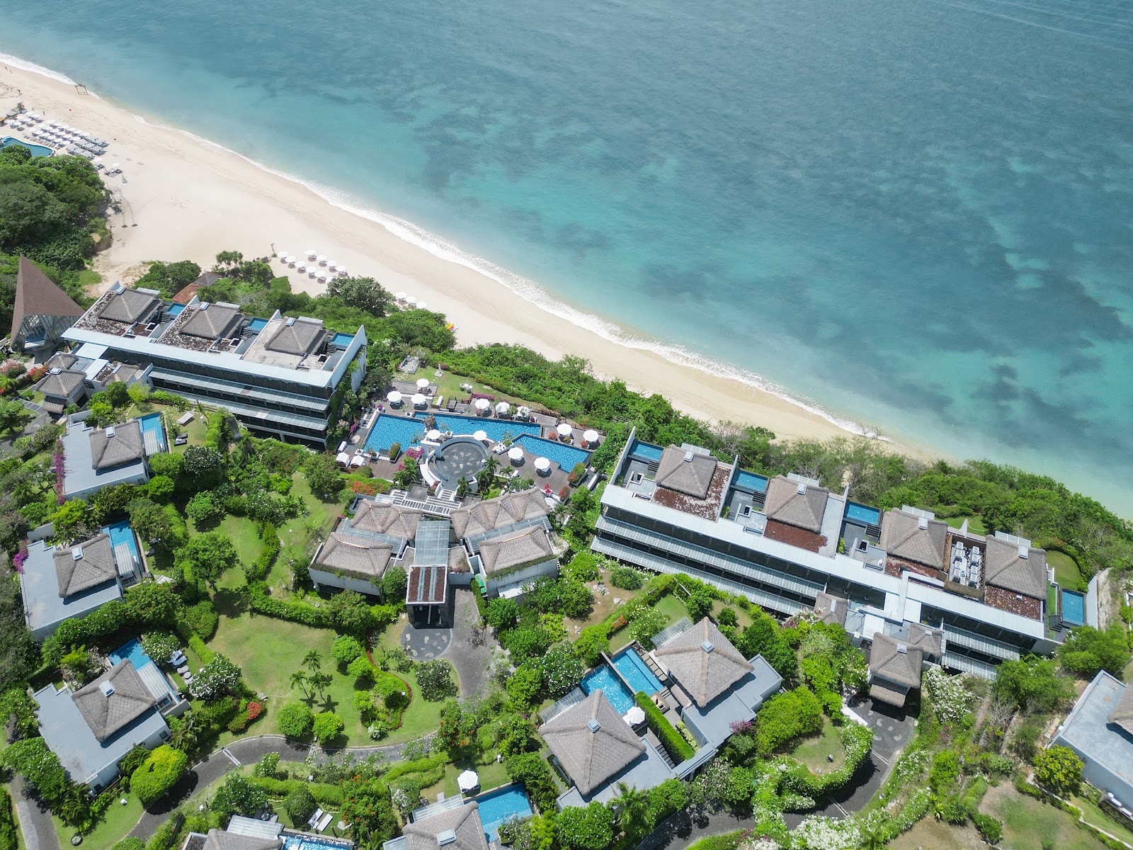 Aerial view of Samabe Bali Suites & Villas with private villas, pools, and central courtyard nestled between lush grounds and turquoise ocean beach.