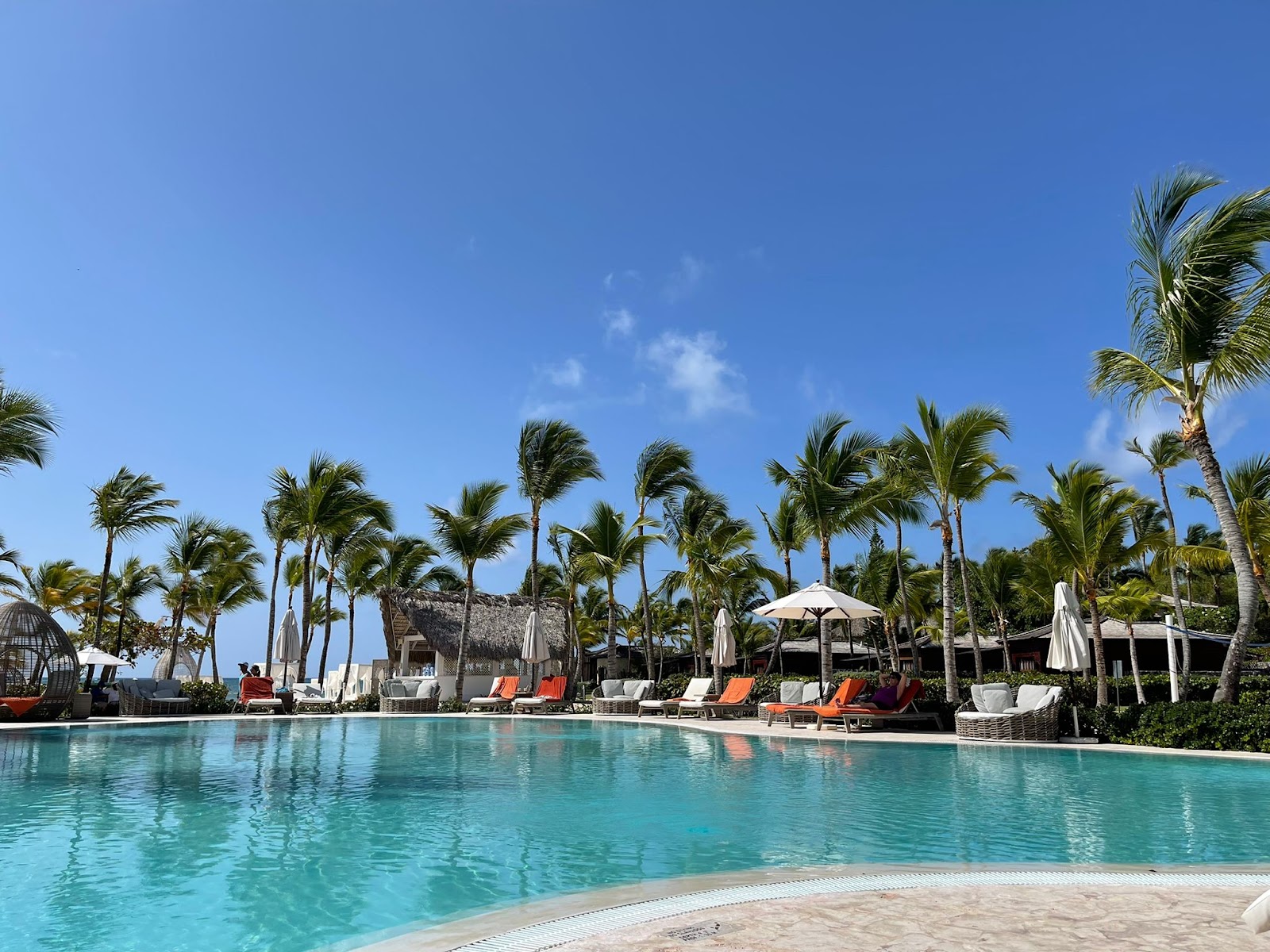 Turquoise pool with sandy beach in foreground, palm trees lining the deck, white and orange lounge chairs, umbrellas, and thatched-roof structures at Sanctuary Cap Cana.