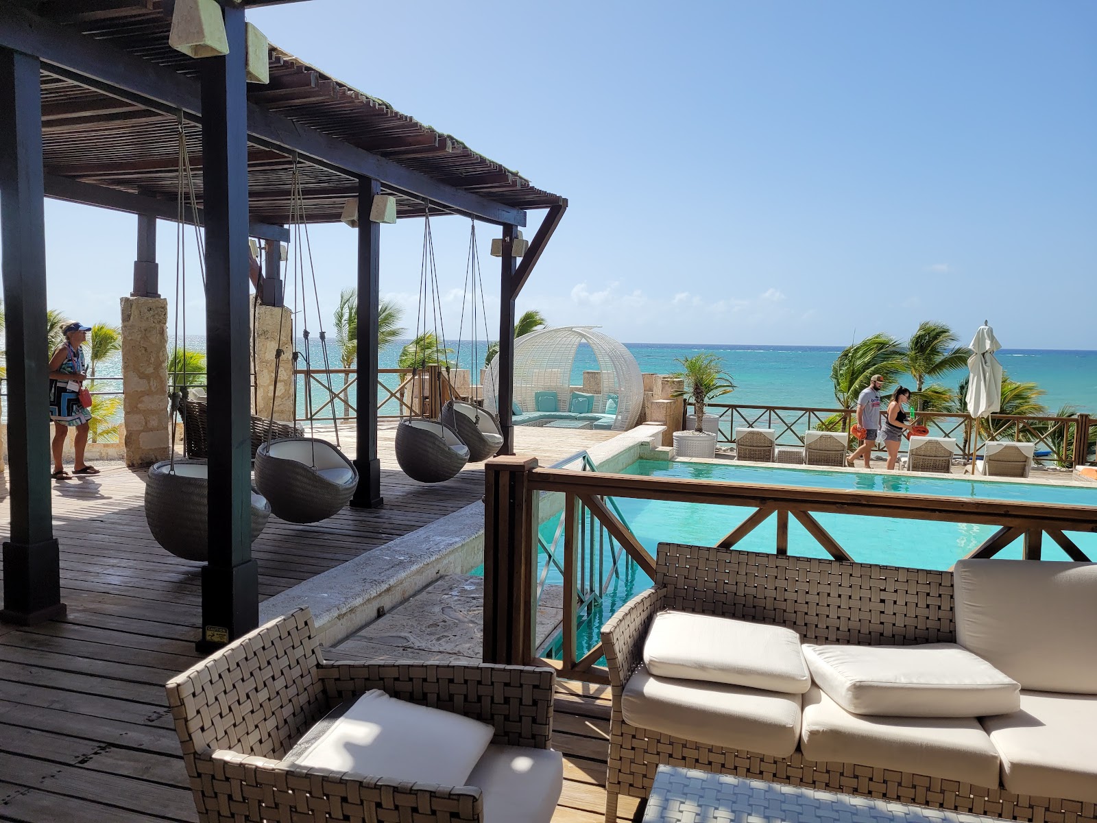 Woven lounge chairs with white cushions overlook a turquoise pool and Caribbean Sea at Sanctuary Cap Cana, with hanging swings and a geodesic dome spa structure visible.