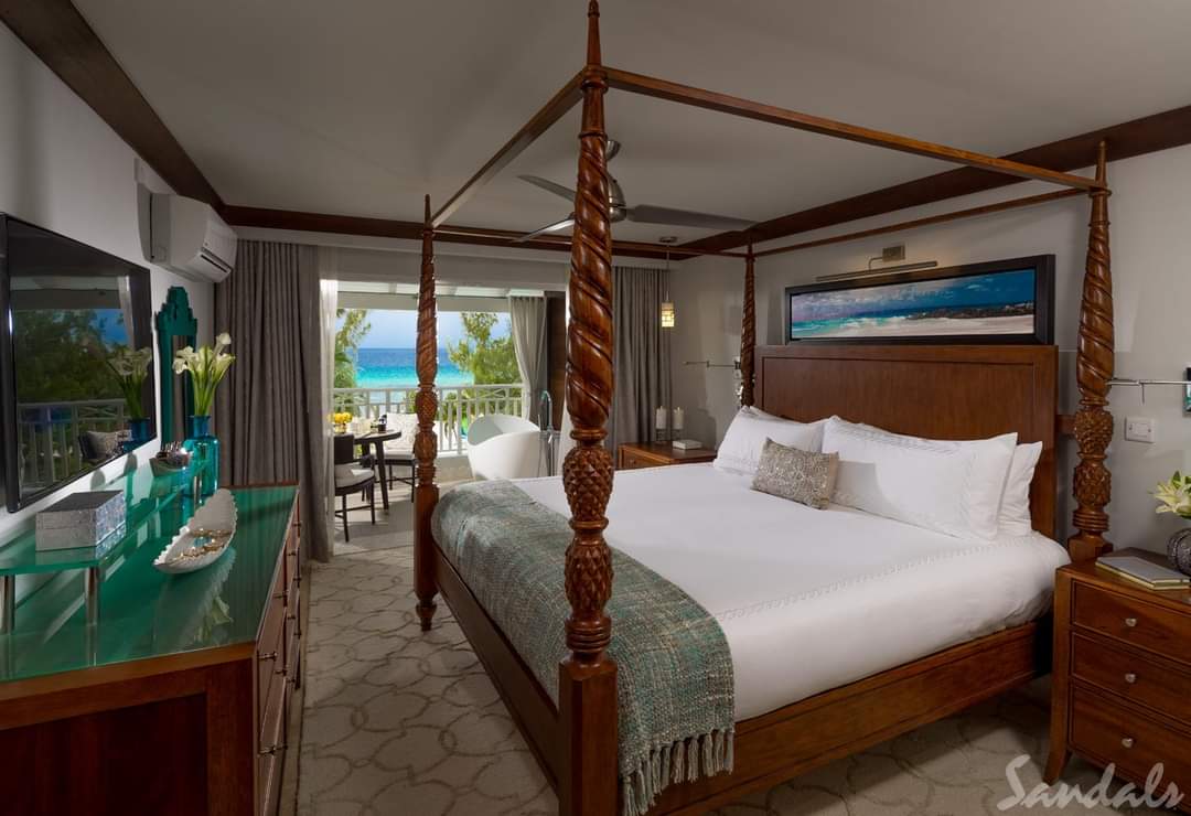Sandals Barbados oceanview suite with four-poster bed, green vanity, and balcony overlooking turquoise waters and beach.