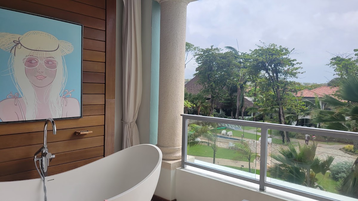 Freestanding bathtub beside a balcony overlooking tropical grounds at Sandals Barbados, with pop art portrait on wood-paneled wall.