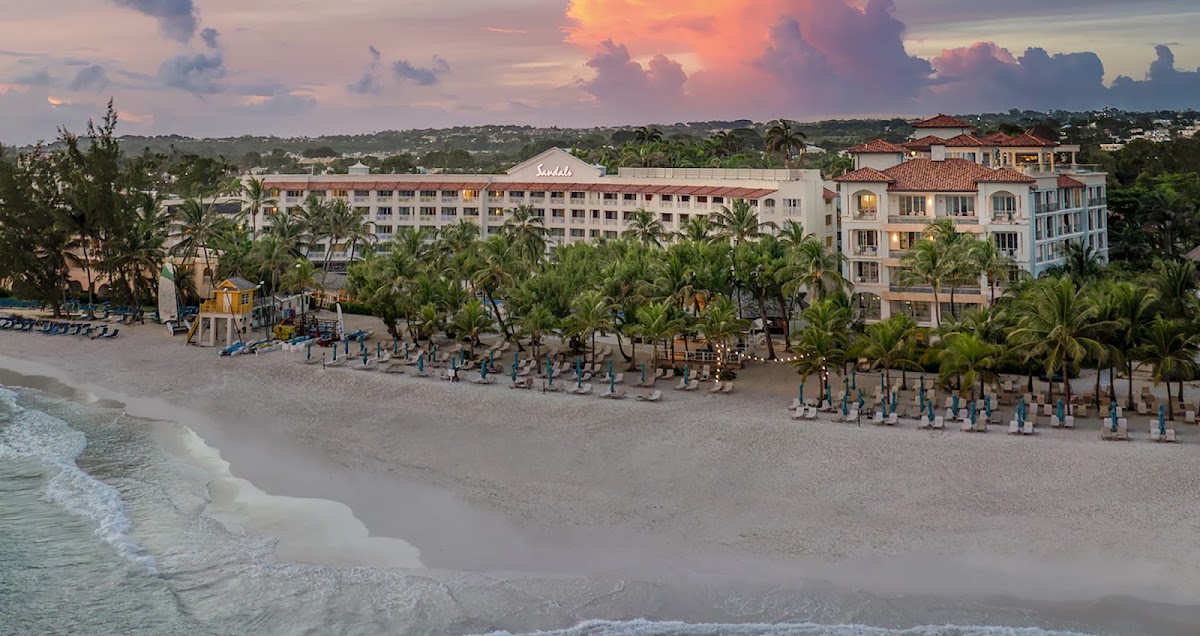 Sandals Barbados beachfront at sunset with pink and coral-colored buildings, palm trees, beach loungers, and gentle waves rolling onto white sand.