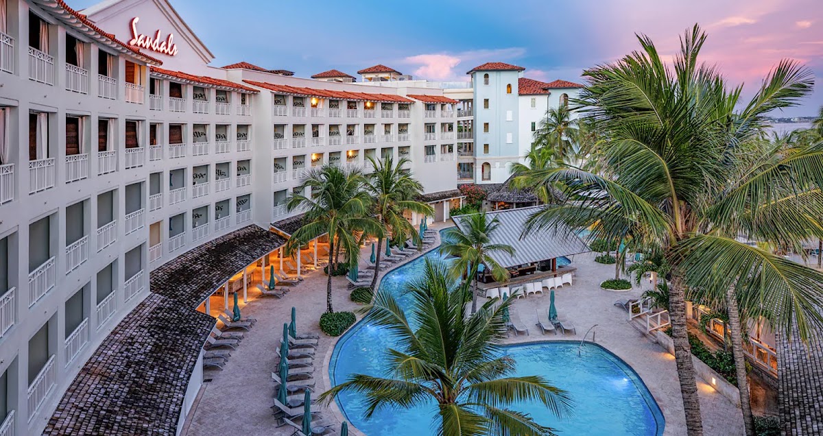 Sandals Barbados courtyard at dusk with white colonial-style buildings, curved pool, palm trees, and lounge chairs surrounding the deck.