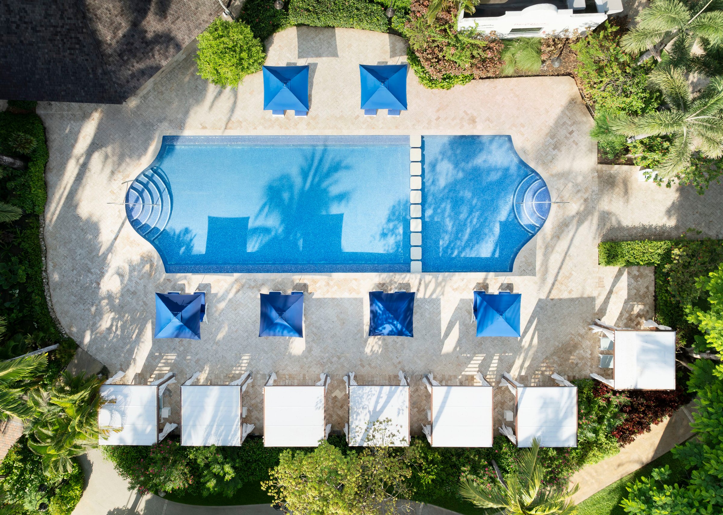 Top-down aerial of a pool with blue umbrellas and white cabanas.