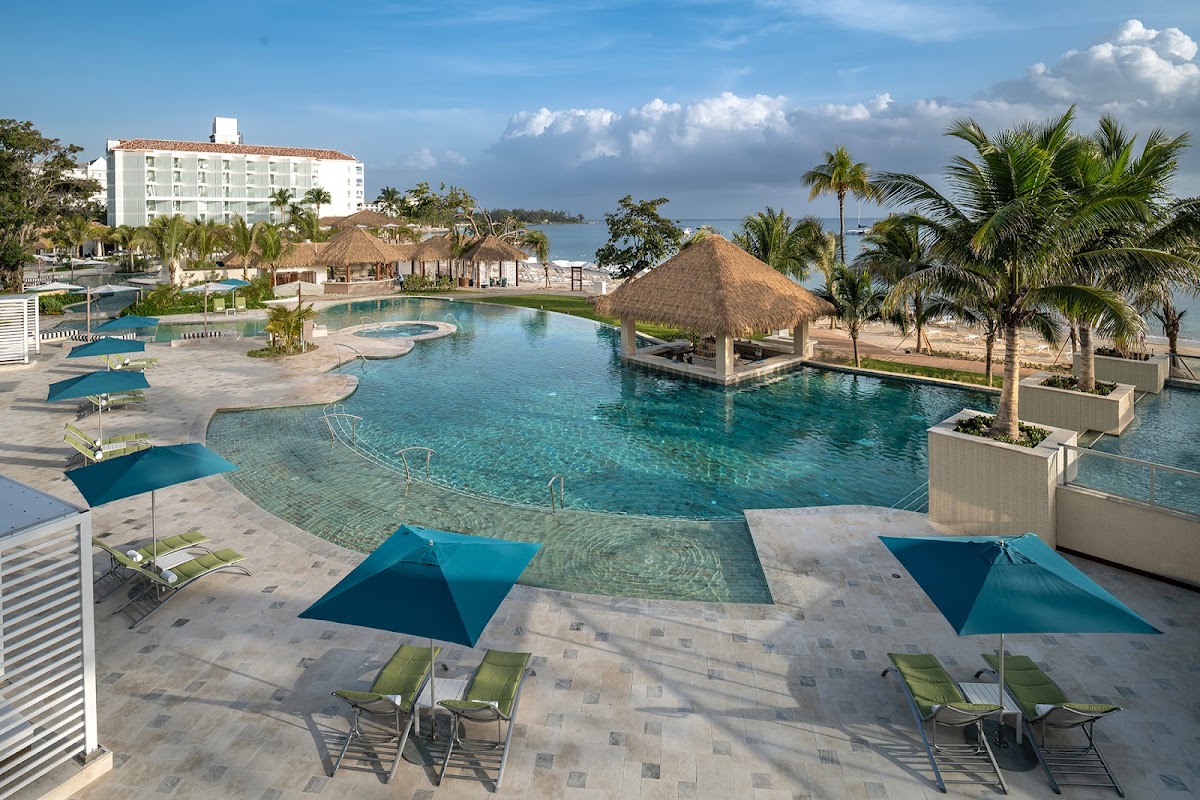 Sandals Dunn's River lagoon pool with thatched-roof cabanas, blue umbrellas, lounge chairs, and the main hotel building overlooking the Caribbean.