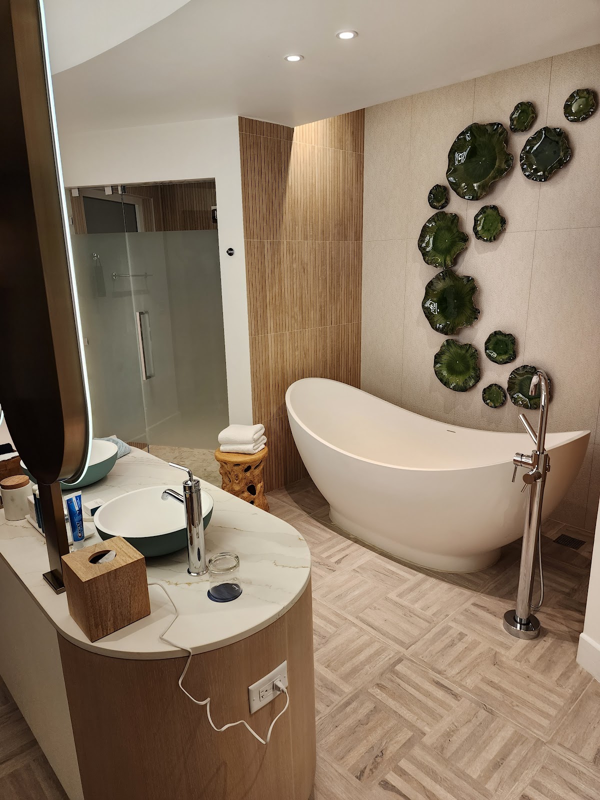 Modern bathroom with freestanding white tub, glass shower enclosure, round vanity sink, and decorative green ceramic plates on the wall.