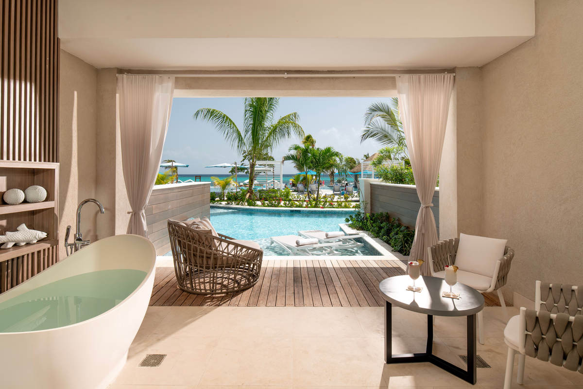 Luxury resort room with oval soaking tub, open to a wooden deck overlooking a lagoon pool, palm trees, and Caribbean Sea beyond.