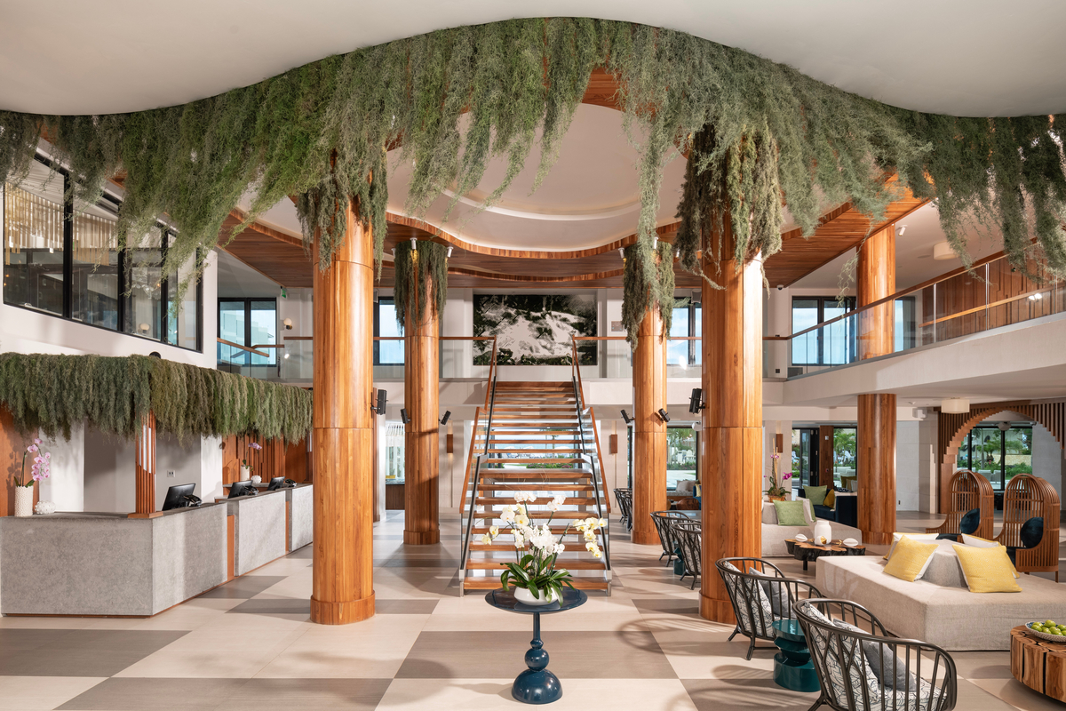 Modern lobby with wooden columns, hanging green plants, central staircase, and seating areas at Sandals Dunn's River resort.