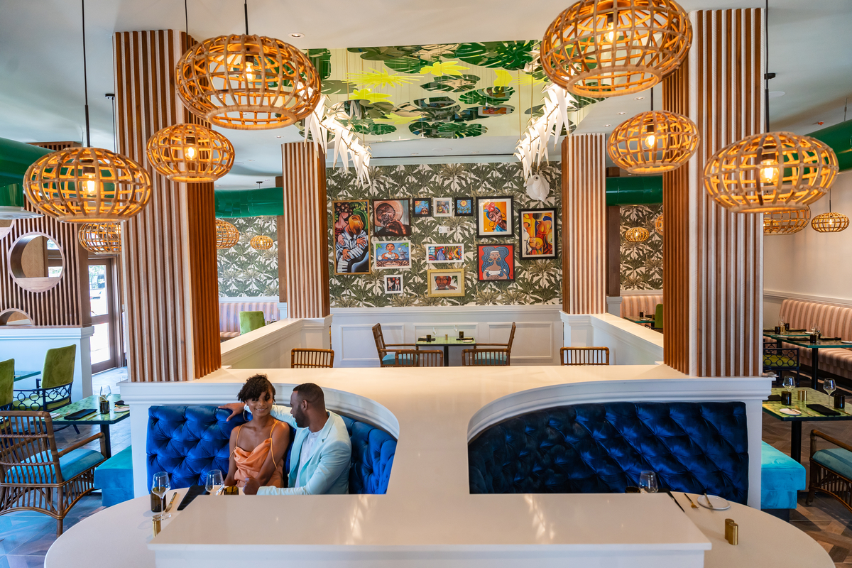 Sandals Dunn's River bar interior with couple seated in blue velvet booth beneath woven globe pendant lights and botanical artwork.