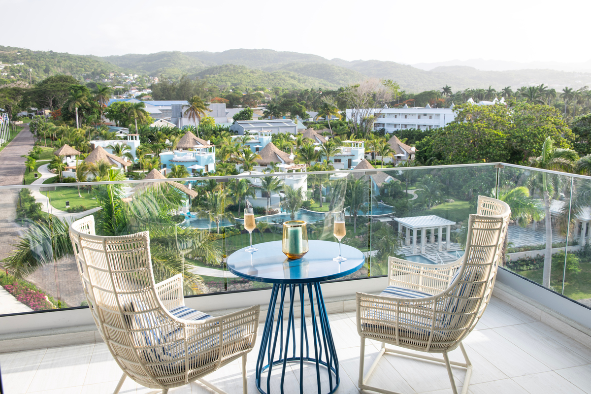 Two wicker chairs face a small blue table with champagne glasses on a balcony overlooking Sandals Dunn's River resort, pools, and tropical mountains.