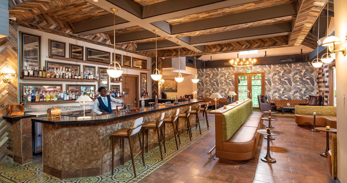 Bar and lounge area with wooden beamed ceiling, curved bar counter, hanging pendant lights, and seating with green upholstery at Sandals Dunn's River.