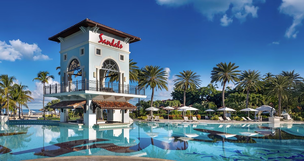 Sandals Grande Antigua's beachfront pool complex with a distinctive white tower, palm trees, and lounge seating under umbrellas beneath blue sky.