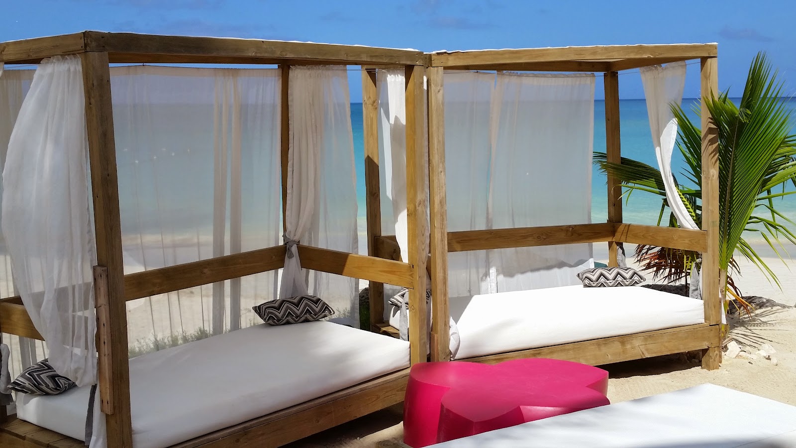 Wooden cabana beds with white curtains and cushions sit on white sand facing turquoise ocean and palm trees under clear blue sky.