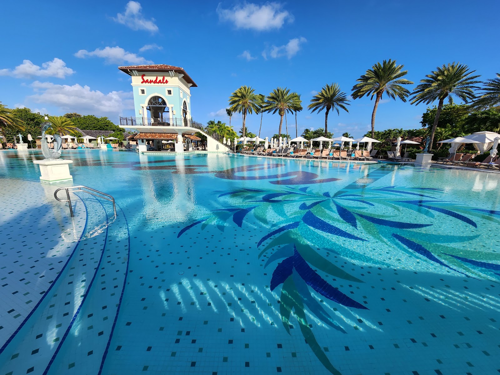 Large turquoise pool with decorative mosaic design at Sandals Grande Antigua, white tower and palm trees visible under blue sky.