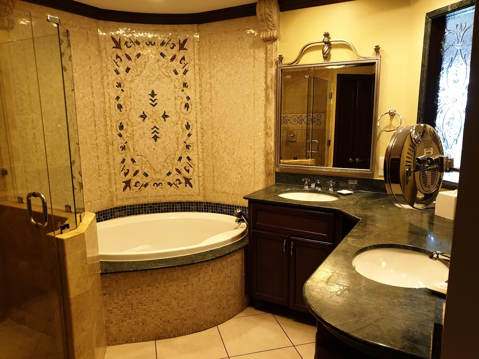 Luxury bathroom with curved soaking tub, dual vanities, walk-in shower, and gold-toned fixtures and decorative wall tile work.