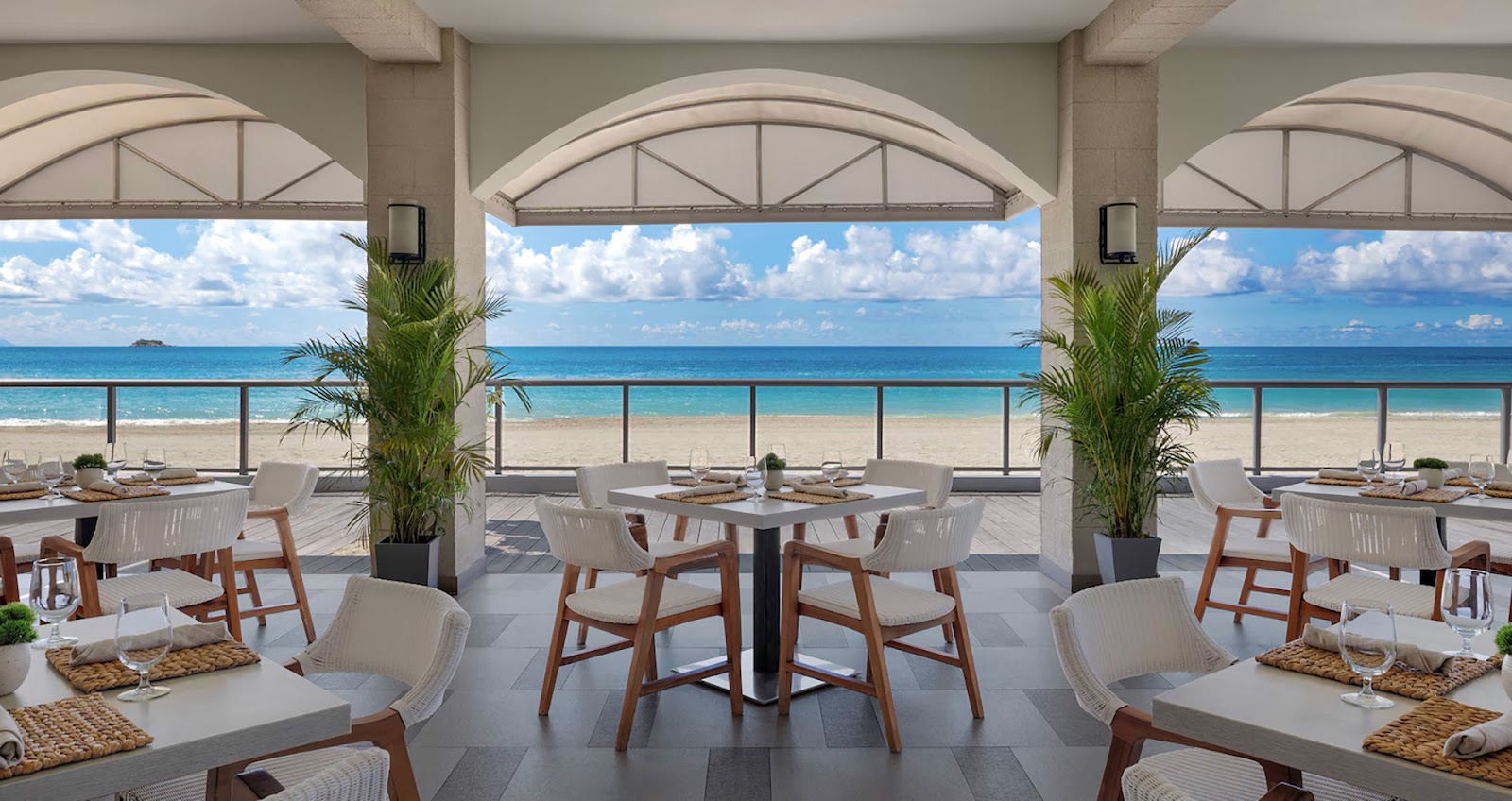 Beachfront dining pavilion at Sandals Grande Antigua with arched shade structures, wooden chairs, and turquoise ocean views beyond the sand.