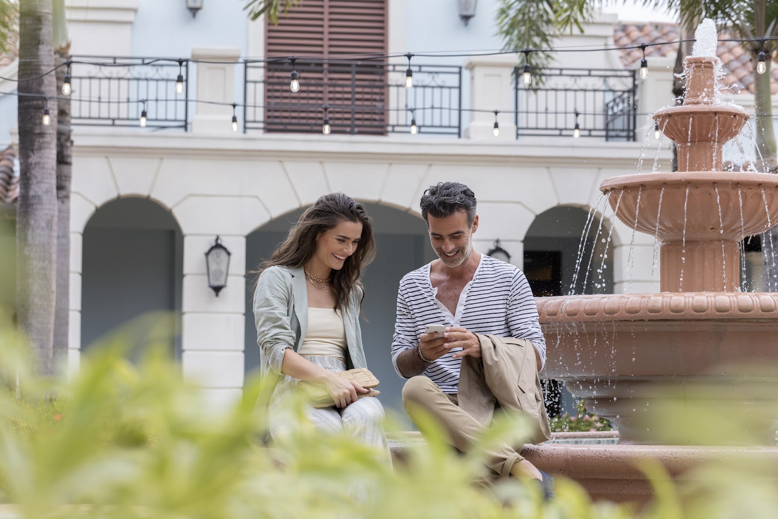 A couple sits by a tiered fountain in a courtyard with white colonial architecture, arched doorways, and a railed balcony above.