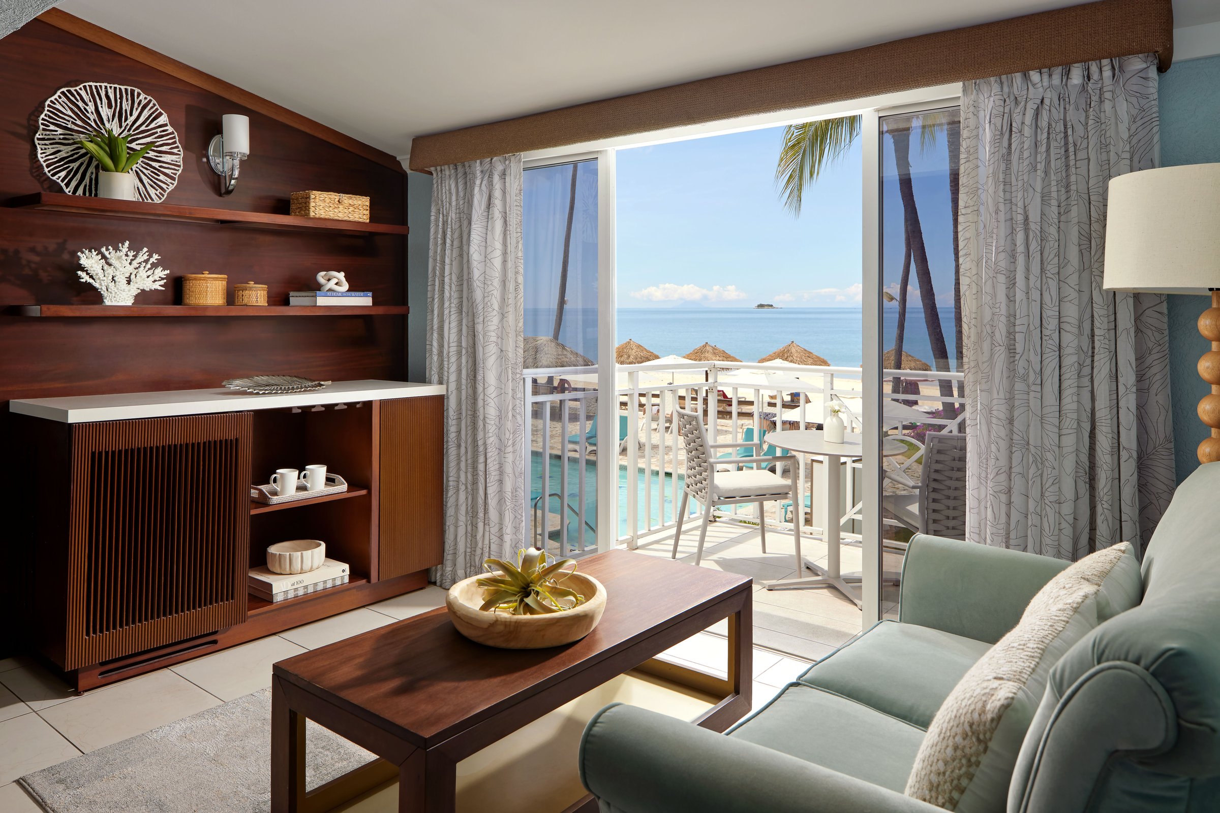 Sitting area with a sofa and wood credenza by a balcony over the pool and beach.