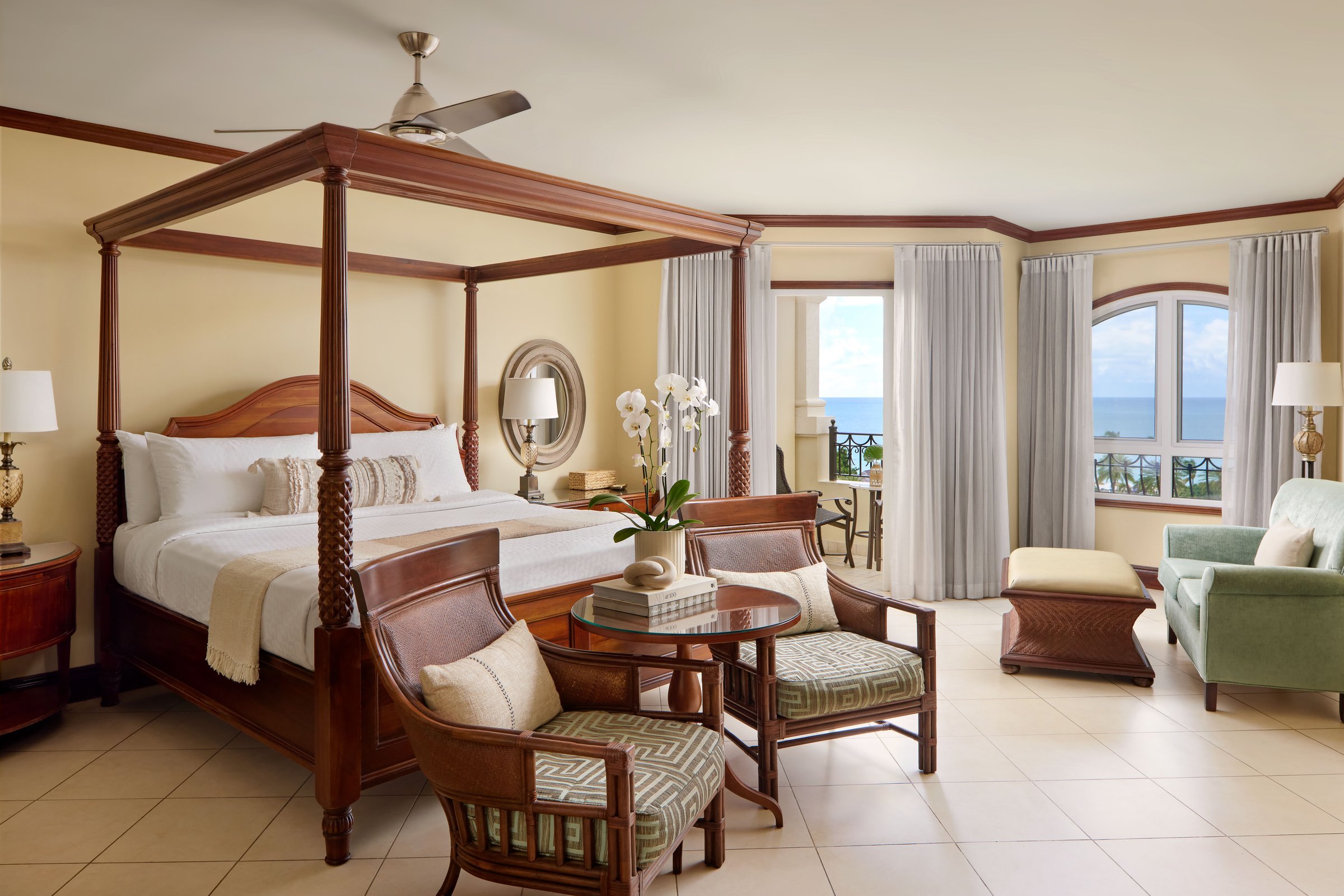 Spacious suite with a four-poster bed, armchairs and arched ocean-view windows.