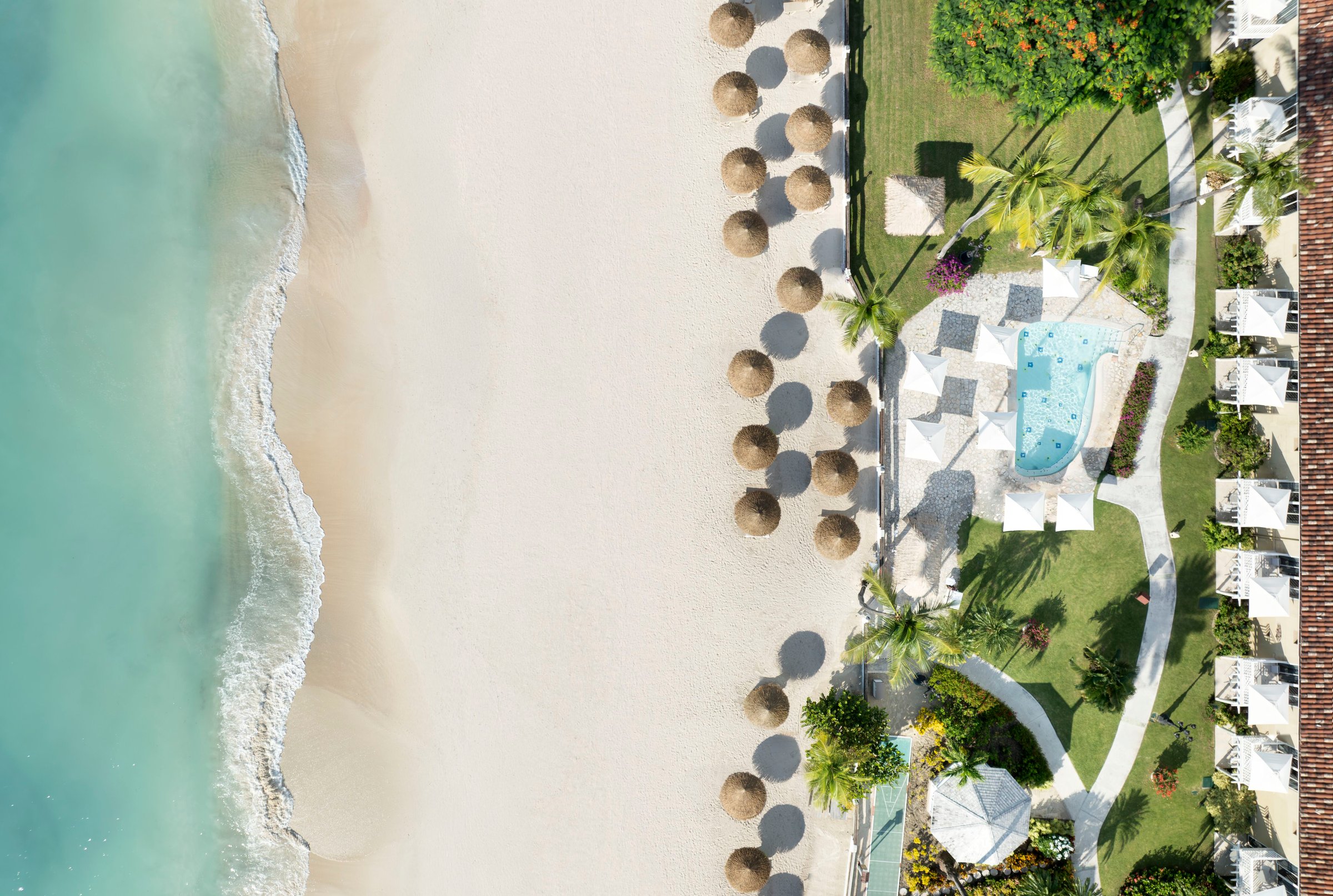 Top-down aerial of the beach, thatched umbrellas and a garden pool behind.