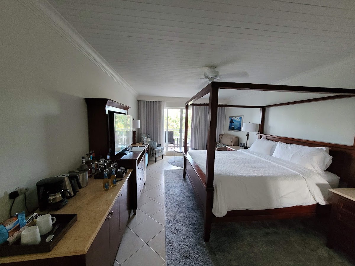 Spacious resort room with four-poster bed, wet bar with coffee maker, and balcony access overlooking tropical grounds.