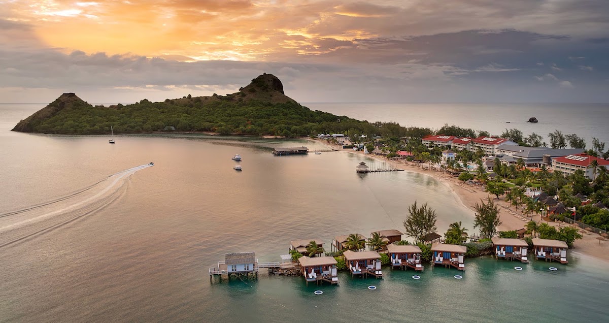 Sandals Grande St. Lucian resort overlooks a bay with overwater bungalows, a sandy beach, palm trees, and a forested island peak at sunset.