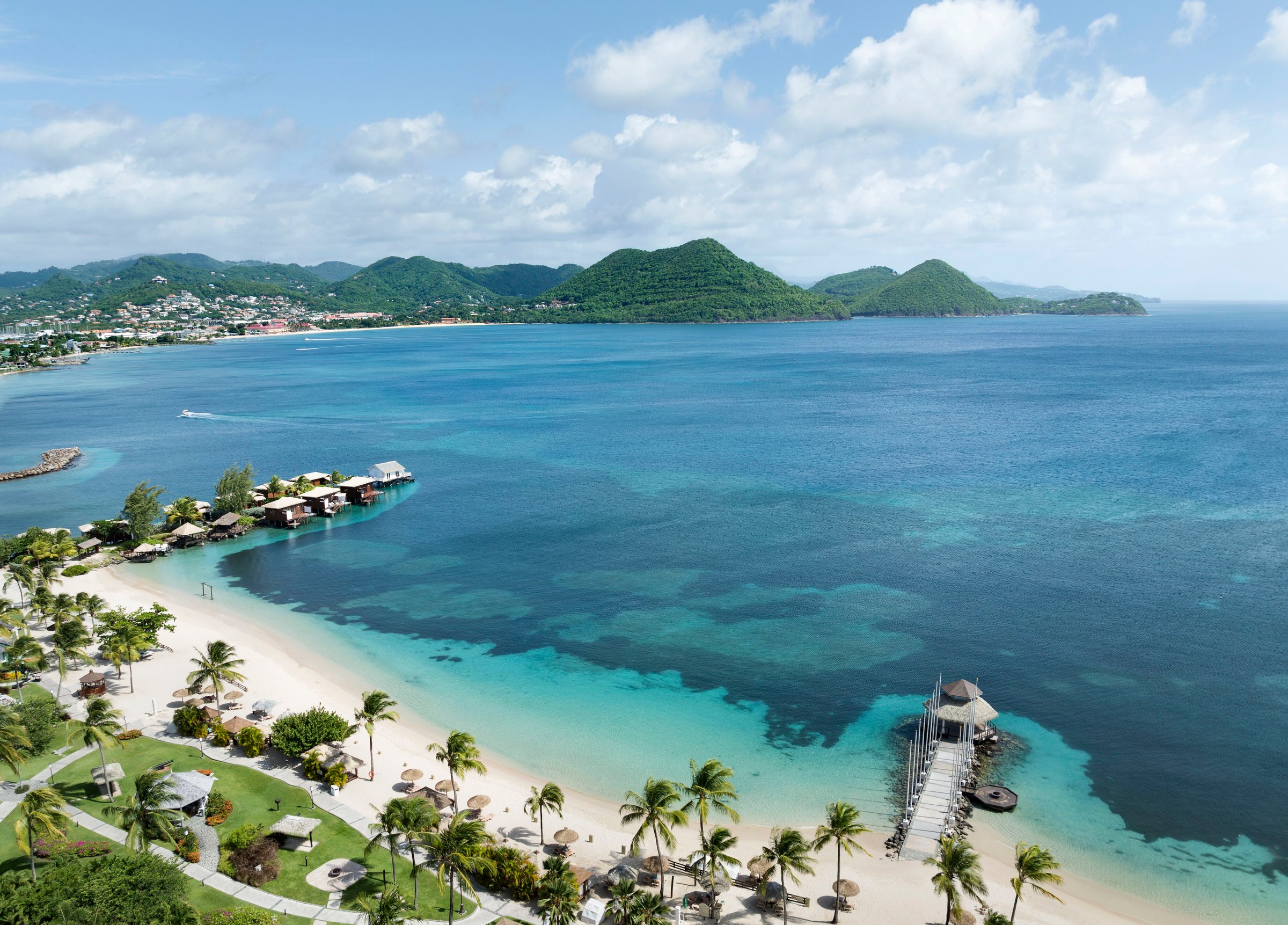 Aerial along the beach to the overwater bungalows with Rodney Bay and green hills behind.