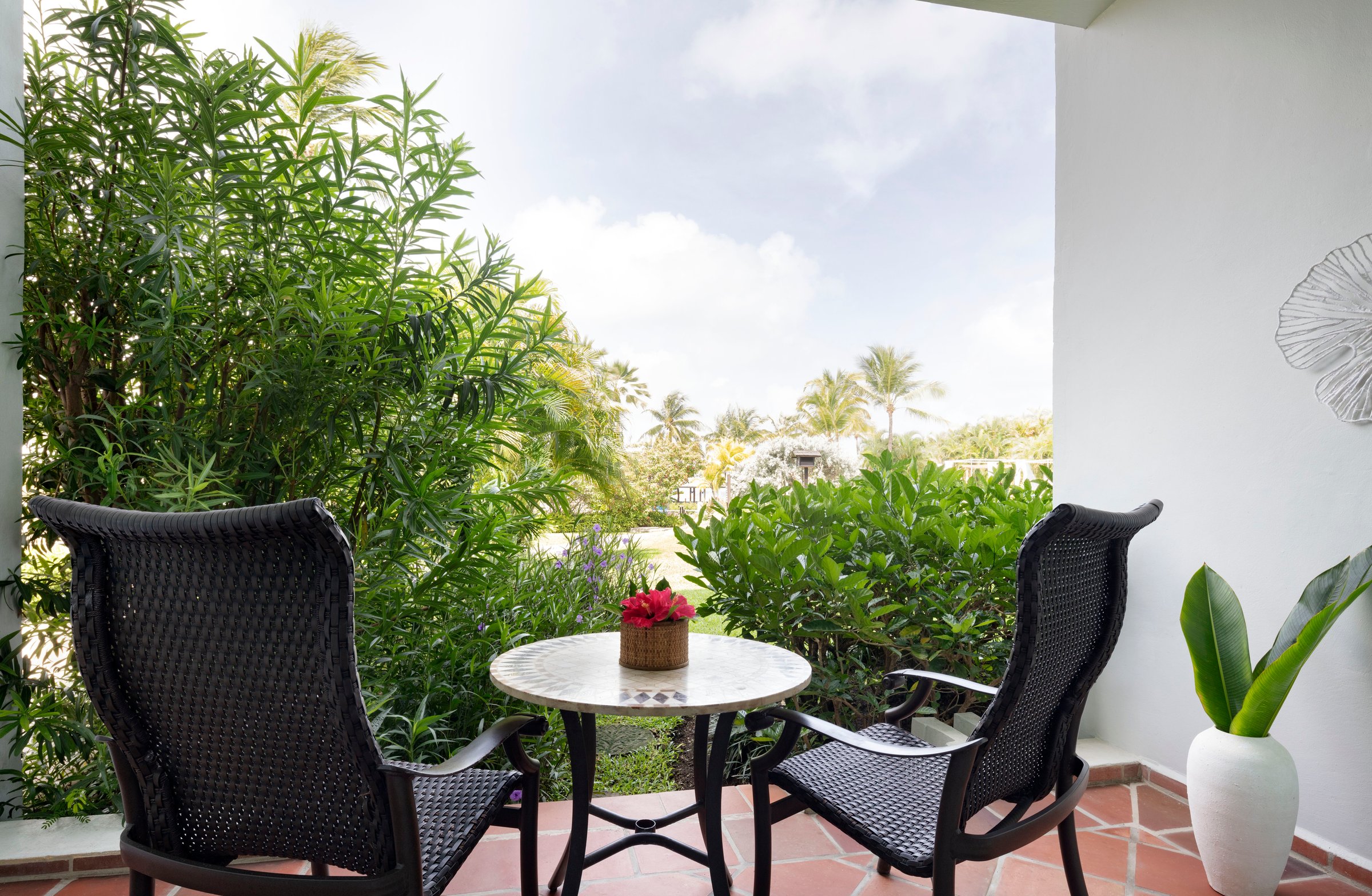 Garden balcony with wicker chairs and a bistro table among greenery.
